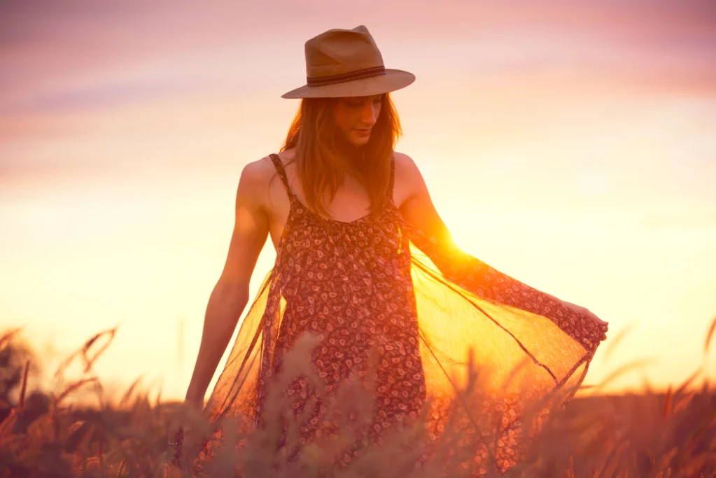 Young woman in a floral dress and hat standing in a field at sunset, bathed in warm light, representing healing, self-awareness, and the journey to heal childhood trauma.