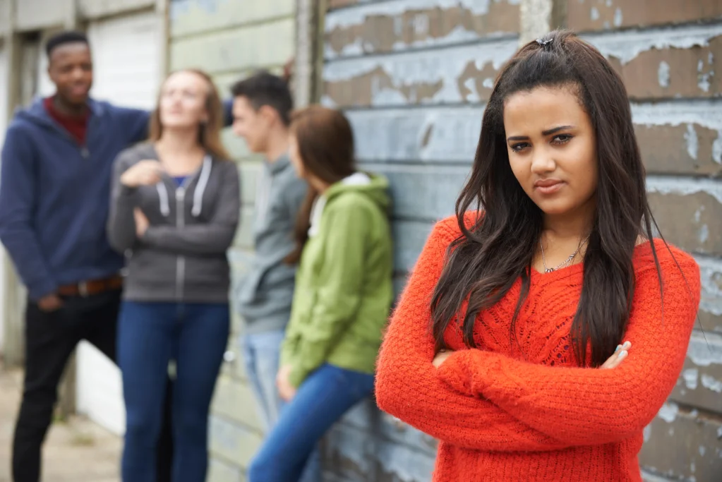 A serious-looking teenage girl with long dark hair and a red sweater stands with arms crossed. In the background, a group of teens chats near a worn-down urban wall—capturing the challenges of parenting today’s teens in a social world.