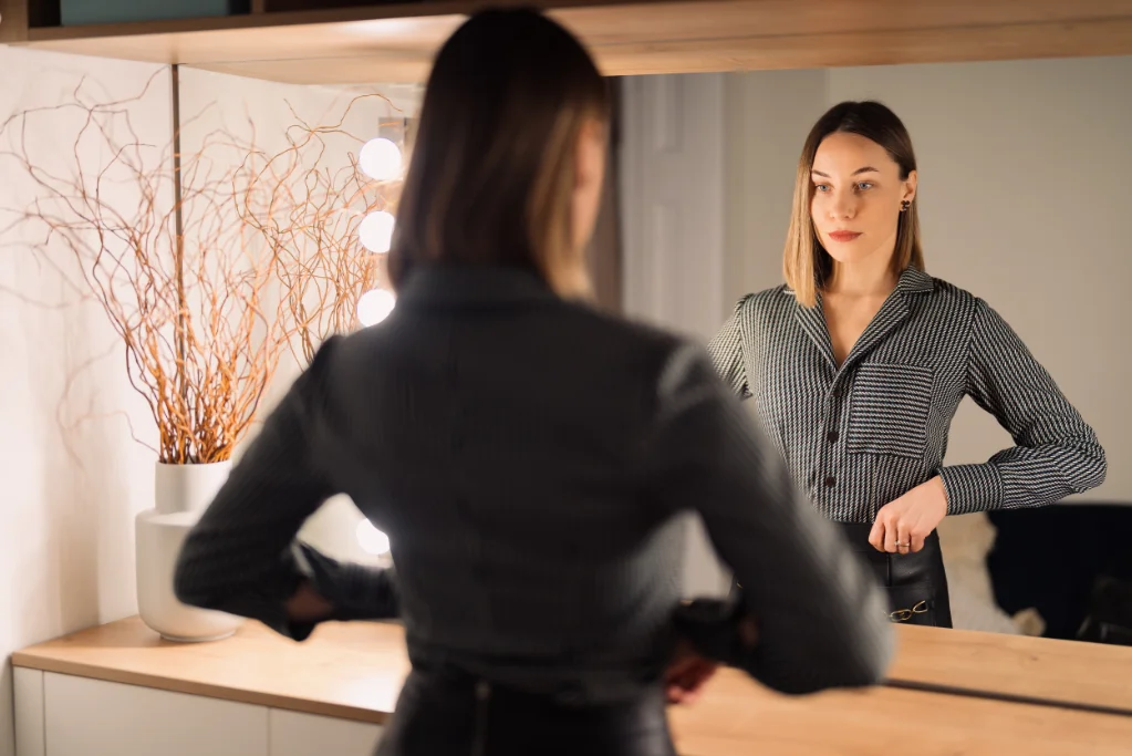 A woman stands in front of a mirror, adjusting her outfit with a thoughtful expression. Her gaze reflects self-reflection, confidence, or quiet self-doubt—symbolizing the journey toward inner awareness and balance.