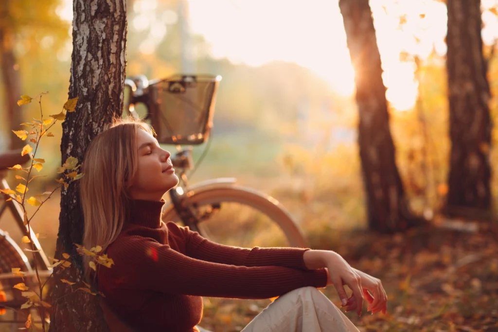Woman sitting peacefully against a tree in autumn sunlight, eyes closed and breathing calmly, symbolizing relaxation and nervous system balance.