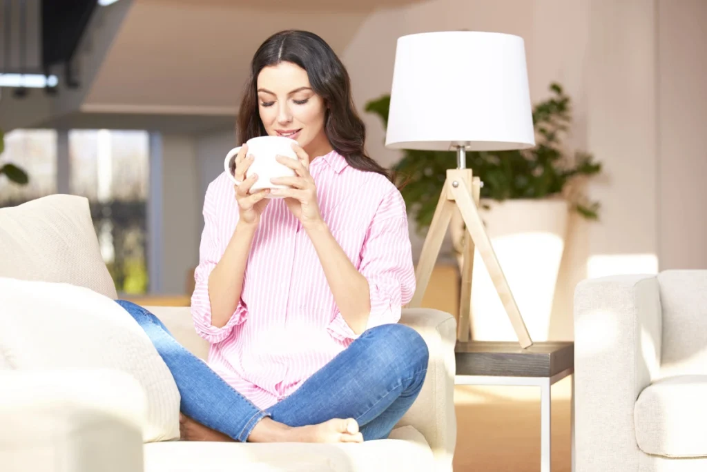 A woman sitting cross-legged on a couch holding a large mug, relaxing in a bright, softly lit living room with a lamp and plants in the background.