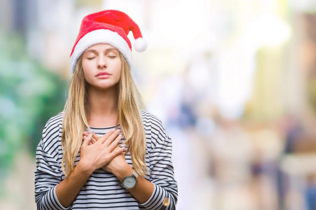 Woman wearing a Santa hat with eyes closed and hands over her chest, pausing to breathe calmly in a softly blurred outdoor setting.